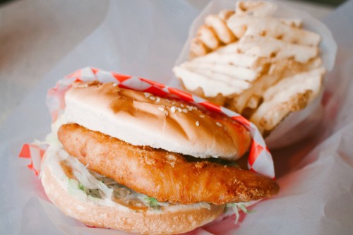 A crispy fish sandwich with lettuce on a sesame seed bun, accompanied by a side of waffle fries.