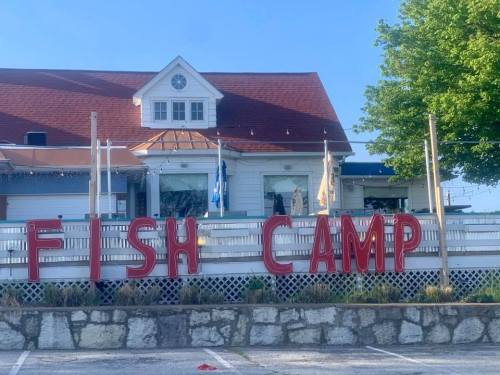 A building with a red roof and a sign that reads "FISH CAMP" in large letters, surrounded by greenery.