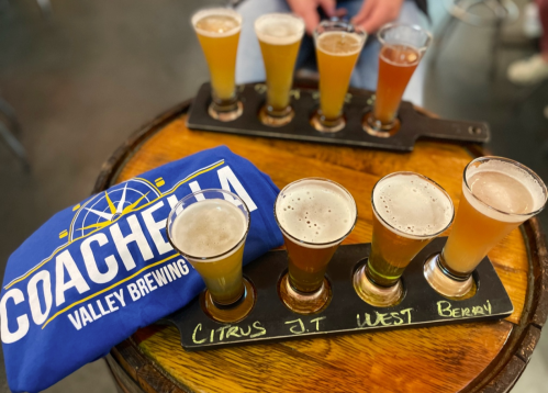 A wooden tray with six small beer glasses, a blue Coachella Valley Brewing shirt, and a wooden table background.
