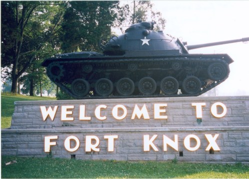 A tank on display beside a sign that reads "Welcome to Fort Knox," surrounded by trees and grass.