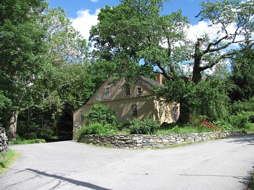 A quaint yellow house surrounded by trees, situated at the intersection of two roads on a sunny day.