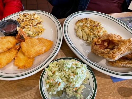 Two plates of food featuring fried shrimp, crab cakes, fried chicken, and a side of coleslaw.