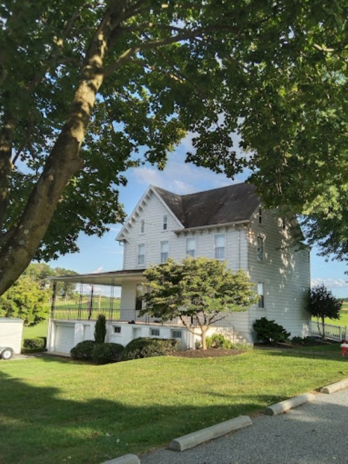 A white two-story house surrounded by greenery and trees, with a porch and a well-maintained lawn.