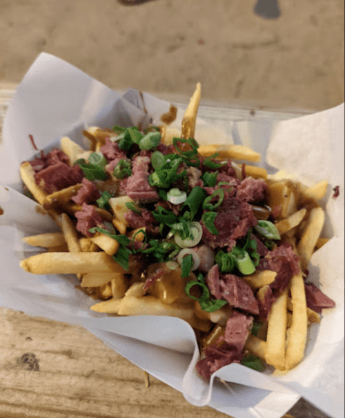 A serving of French fries topped with chopped meat and green onions, served in a paper tray.