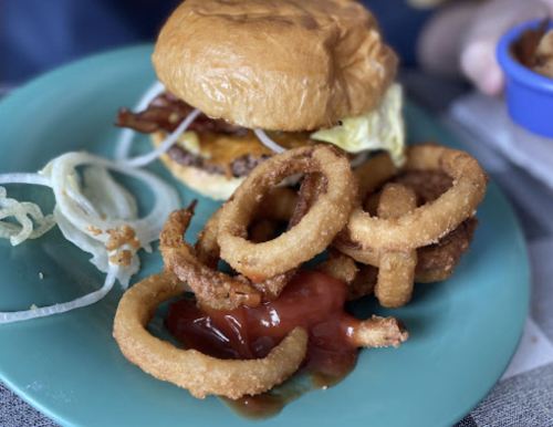 A burger with lettuce, onion, and bacon on a plate, accompanied by crispy onion rings and ketchup.