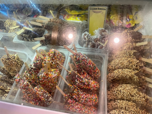 A display of frozen bananas coated in chocolate and toppings, including sprinkles and nuts, in a glass case.