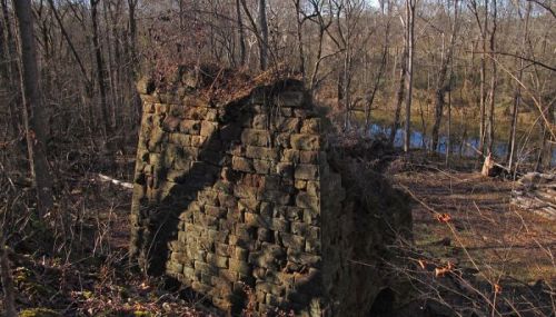 A weathered stone wall covered in moss, surrounded by bare trees and a glimpse of a pond in the background.