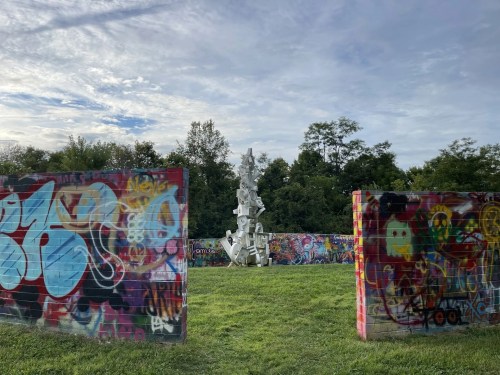 A white sculpture stands amidst colorful graffiti-covered walls in a grassy area under a cloudy sky.