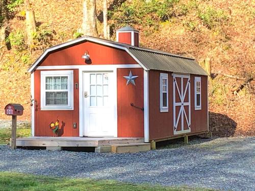 A small red barn-style cabin with a white door and star decoration, set in a wooded area with gravel in front.