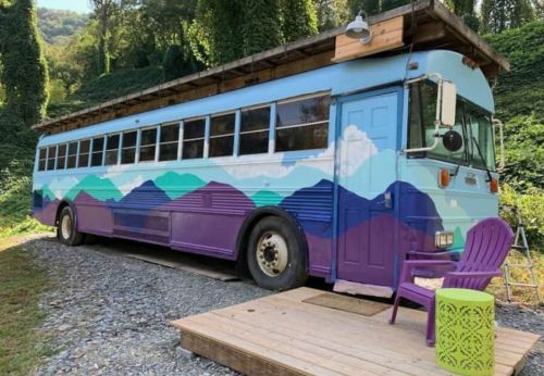 A colorful converted bus with mountain murals parked beside a wooden deck and a purple chair. Lush greenery surrounds it.