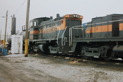 A vintage diesel locomotive with "HARBOR" painted on the side, parked near industrial buildings and gravel.