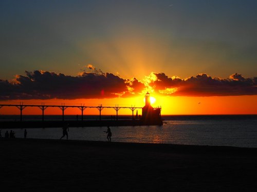 Silhouette of a lighthouse at sunset, with rays of light and clouds over a calm sea and people walking on the beach.