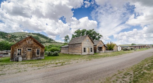 A dusty road lined with rustic wooden houses under a blue sky with fluffy clouds and green hills in the background.