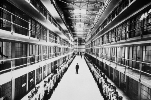 A black and white photo of a prison interior, with rows of cells and guards lined up facing a central figure.