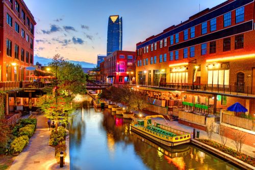 A scenic view of a canal lined with buildings, boats, and trees, with a city skyline at sunset in the background.