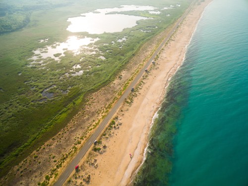 Aerial view of a sandy beach along a turquoise sea, with lush green wetlands and ponds in the background.