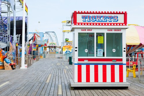 A colorful ticket booth on a wooden boardwalk, with amusement rides in the background under a clear sky.