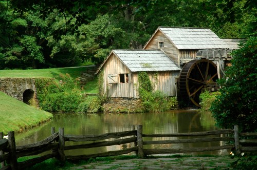 A rustic wooden mill with a water wheel beside a calm pond, surrounded by lush greenery and a wooden fence.