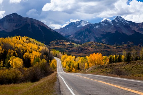 A winding road through vibrant autumn trees, with majestic mountains and a cloudy sky in the background.