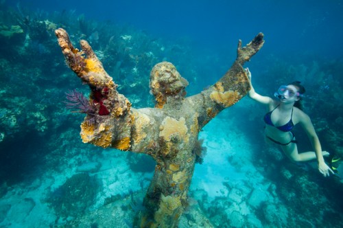 A diver swims near a submerged statue of Christ, covered in coral, in a clear blue ocean.