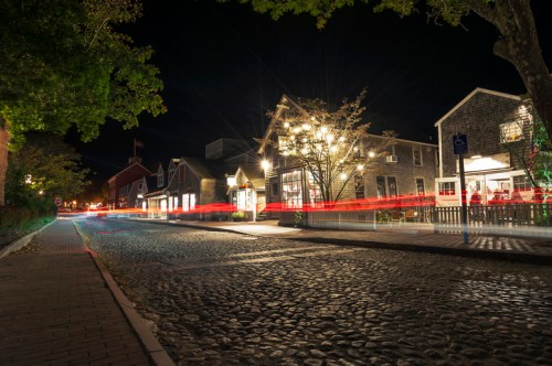 A cobblestone street at night, lined with buildings, illuminated by lights and streaks from passing vehicles.