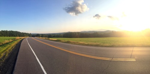 A winding road stretches through a green field, with mountains in the distance and a sunset casting warm light.