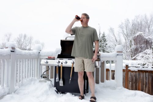 A man in shorts and a t-shirt drinks a beer while standing by a grill in a snowy backyard.