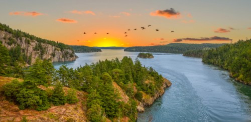 A serene sunset over a river, surrounded by lush green trees and cliffs, with birds flying across the sky.