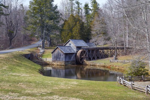 A rustic wooden mill with a water wheel beside a calm pond, surrounded by trees and a winding road.