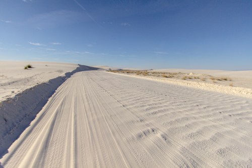 A sandy road stretches through a vast, white desert landscape under a clear blue sky.