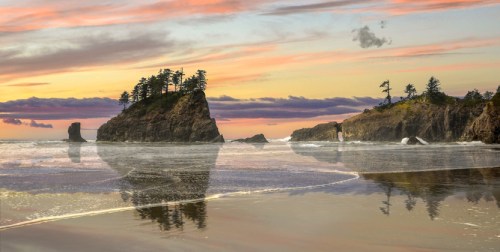A serene beach scene at sunset, featuring rocky outcrops and trees reflected in the wet sand.