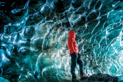 A person in a red hoodie stands in an ice cave, surrounded by shimmering blue and clear ice formations.