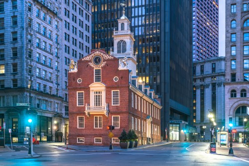 Historic brick building with a clock tower, surrounded by modern skyscrapers in a city setting at dusk.