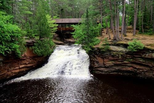 A waterfall cascades over rocky cliffs, surrounded by lush green trees and a wooden structure above.