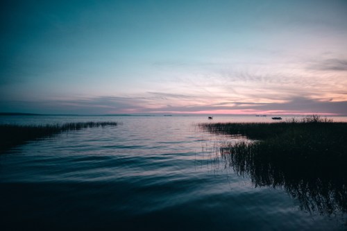 A serene lake at sunset, with calm waters reflecting a colorful sky and lush grass along the shoreline.