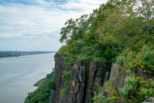 A scenic view of a rocky cliff with lush greenery overlooking a river and distant city skyline under a cloudy sky.