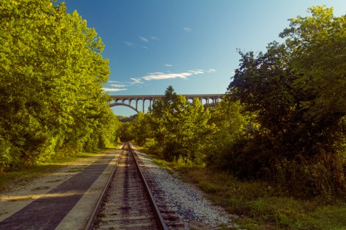 A scenic view of a railway track leading towards a tall bridge, surrounded by lush green trees under a clear blue sky.
