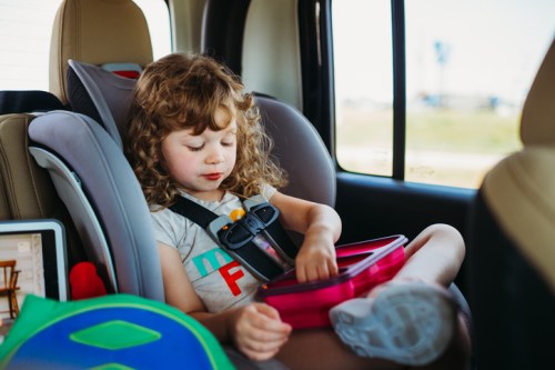 A young child sits in a car seat, focused on a pink lunchbox, with a tablet nearby. Sunlight streams through the window.