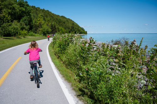 A child rides a bicycle along a scenic road by the water, surrounded by greenery and a clear blue sky.