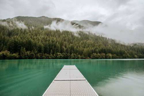 A serene lake surrounded by misty mountains and dense evergreen forests, with a wooden dock extending into the water.
