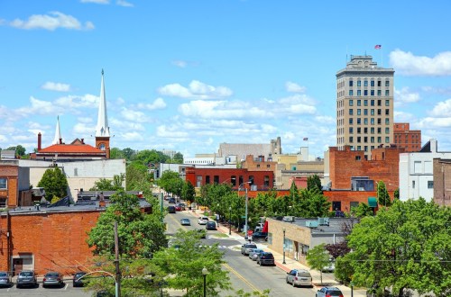 A vibrant cityscape featuring brick buildings, church spires, and a tall tower under a blue sky with fluffy clouds.