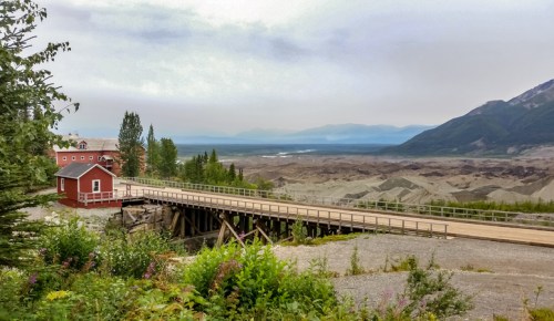 A red building on a wooden bridge overlooks a vast, rugged landscape with mountains and a cloudy sky in the background.