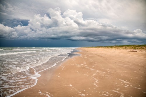 A sandy beach with gentle waves, under a dramatic sky filled with dark clouds.