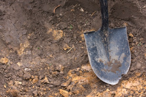 A black shovel resting on freshly dug, dark brown soil with scattered rocks and dirt clumps.