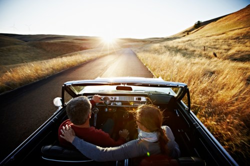 A couple enjoys a scenic drive in a convertible, with golden fields and a sunset in the background.