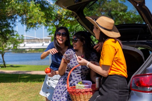 Three friends enjoying a picnic by a lake, laughing and sharing food from a basket while sitting by a car.