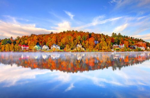 Colorful autumn trees reflect in a calm lake, with charming houses lining the shore under a bright blue sky.