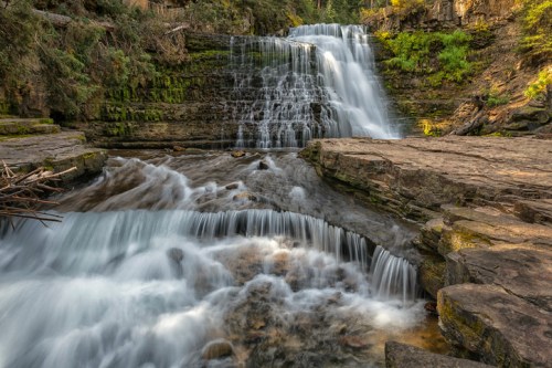 A serene waterfall cascades over rocky ledges, surrounded by lush greenery and smooth flowing water.