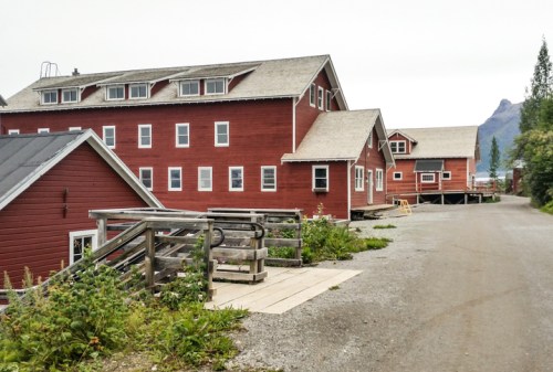 Red wooden buildings line a gravel path, surrounded by greenery and mountains in the background.