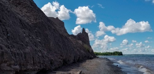 A rocky shoreline under a blue sky with fluffy clouds, featuring steep cliffs and calm water.